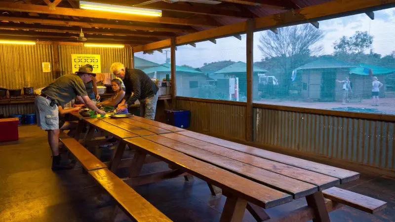 Four travellers cooking at dusk in a rustic outback kitchen on a 3 Day Uluru Adventure tour from Alice Springs, Australia.