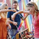 Three friends browsing vibrant handmade crafts and smiling at the lively Chill Out At Byron Bay outdoor market festival.