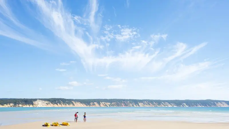 Explore Noosa’s stunning coastline as two adventurers stroll along a sandy beach beside bright yellow kayaks on a Dolphin Sea Kayak 4X4 Tour.