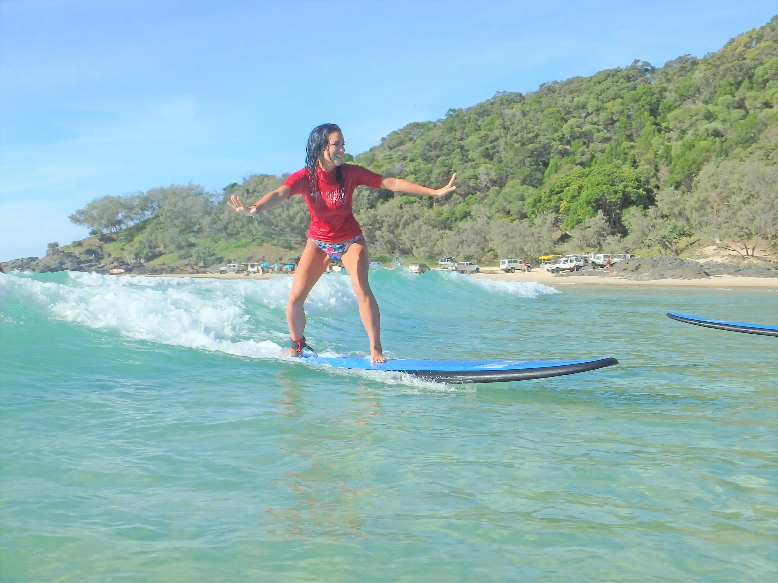 A woman in a vibrant red shirt rides Australia’s Longest Wave by a scenic sandy beach dotted with trees and parked cars in the background.