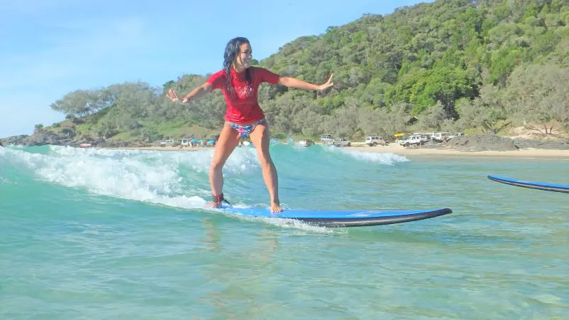 A woman in a vibrant red shirt rides Australia’s Longest Wave by a scenic sandy beach dotted with trees and parked cars in the background.
