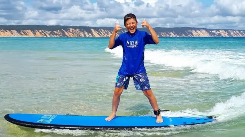 Happy boy giving a thumbs up on a blue surfboard during a Noosa surf lesson in shallow coastal waters, sunny sky in background.