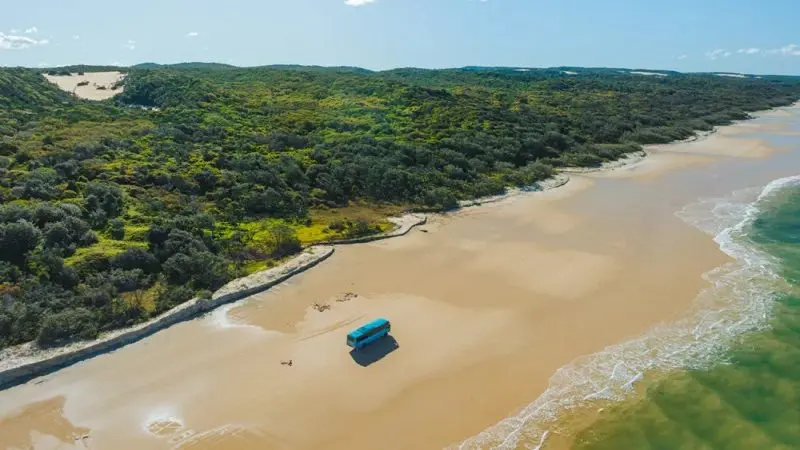 Blue van parked on a vast sandy beach at Kgari 4 Day Experience, framed by lush green forest under clear skies—ultimate adventure scene.