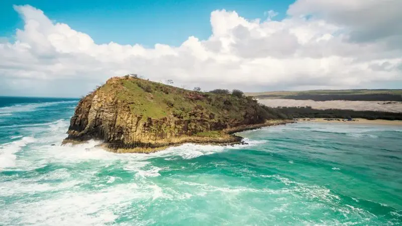 Stunning rocky island with lush greenery, encircled by vibrant turquoise waves—ideal setting for a Kgari 3 Day Getaway beneath clouds.