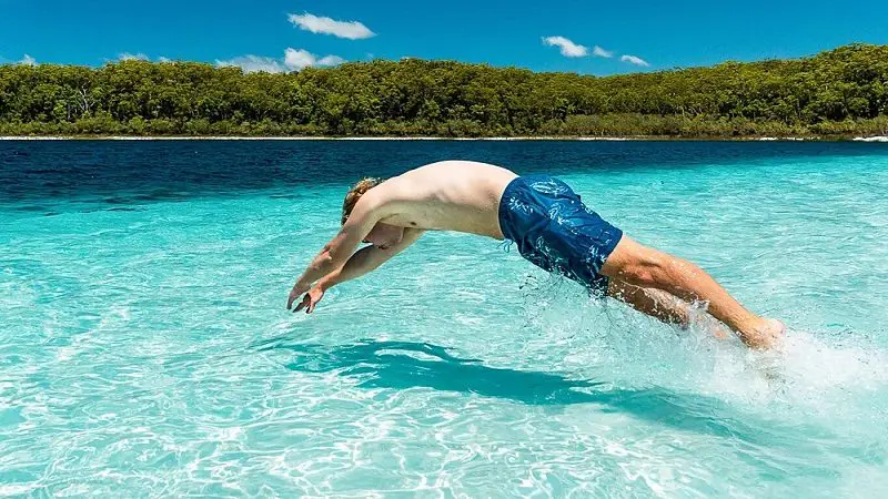 Man wearing blue swimming trunks dives into clear turquoise water on a Kgari 3-Day Getaway beside lush, forested shoreline.