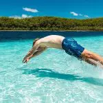 An adventurer in blue swim shorts dives into crystal-clear water by a lush forested shore during a Kgari 3-Day Getaway experience.