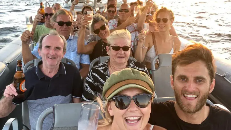 A cheerful group celebrates with drinks on a sunset river cruise, raising glasses and smiling at the camera against scenic views.