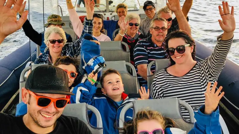 Smiling group enjoying a scenic river cruise, waving hands in the air under bright sunshine, capturing joyful moments on the water.