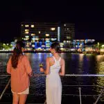 Two women enjoy a night cruise on the Spirit Of Cairns, vibrant harbour lights reflecting as one sips wine onboard the dinner boat.