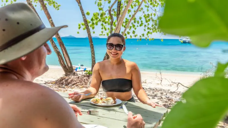 Couple dining at a picnic table on sunlit Frankland Island beach, savouring lunch amid vibrant coastal scenery on Express Tour.
