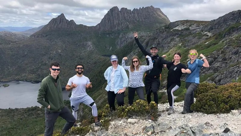 Seven adventurers beam with excitement atop a rugged Cradle Mountain trail on a 2 Day Hobart tour with Wild Tasmania Tours.