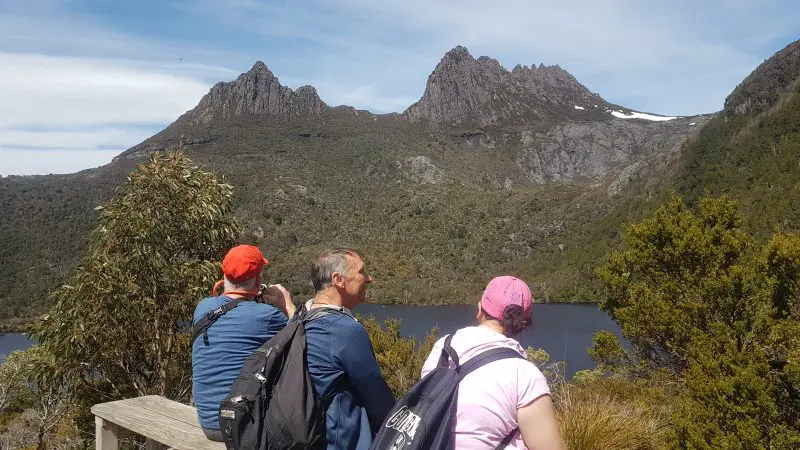 Three hikers with rucksacks rest on a scenic bench, enjoying panoramic wilderness views on the 4 Day Wild Tasmania Wilderness Loop tour.