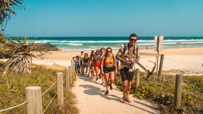 Travellers on the Fully Guided Ultimate East Coast Sydney to Cairns tour walk a sandy path with stunning ocean views and clear blue sky.