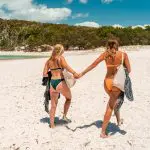 Two women in stylish swimming costumes walk along a sunlit sandy beach on the Fully Guided Ultimate East Coast Sydney to Cairns 3 Week tour.
