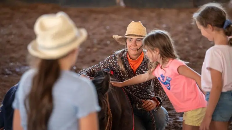 A woman in a cowboy hat kneels beside a horse with three young girls at the 2 Hour Outback Show, capturing Australian rural life.