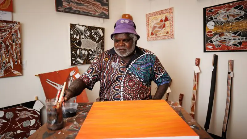 A creative artist in a vibrant purple hat paints on an orange canvas, drawing inspiration from the 4 Day Kakadu Katherine Gorge Experience.