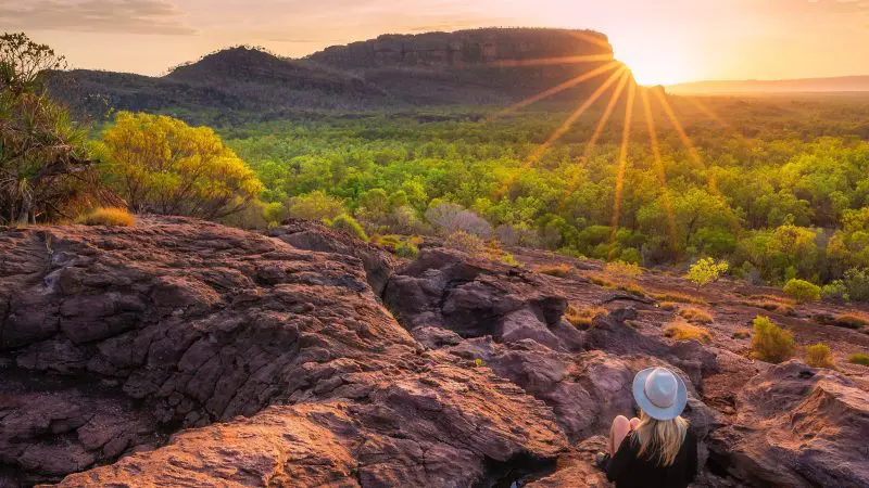 Woman wearing a hat sits on rugged rocks at sunset during a 4-Day Kakadu Katherine Gorge Adventure, enjoying breath-taking views.