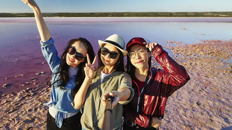 Three friends smile for a selfie beside a vibrant pink lake during their 4-Day Perth to Monkey Mia Coastal Loop Return Tour adventure.