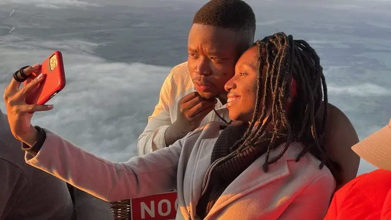 Two people smiling, taking a selfie on a hot air balloon ride with breakfast over Perth’s Avon Valley at sunset, above the clouds.
