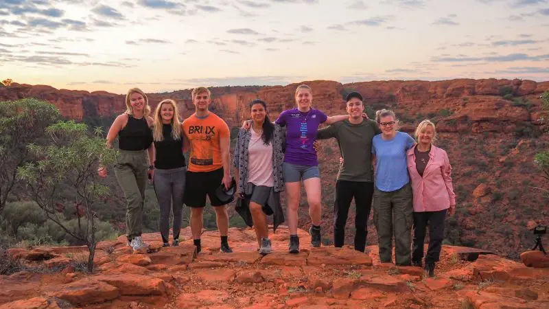 Eight travellers smiling on a scenic rocky cliff at sunset during a 7 Day Uluru to Adelaide tour with Untamed Escapes adventure.
