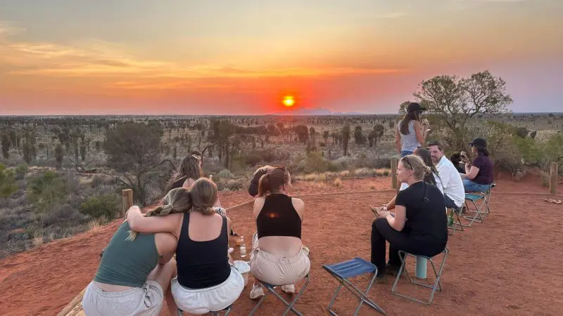 Travellers seated on stools on vibrant red sand admire a stunning sunset during the 7 Day Uluru to Adelaide Untamed Escapes tour.