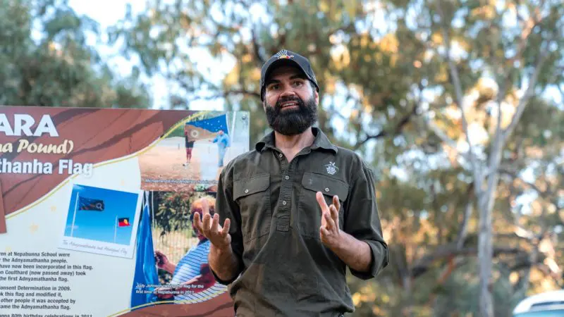 Bearded man sharing insights by the Adnyamathanha Flag during the 7 Day Adelaide to Uluru Adventure tour with Untamed Escapes.