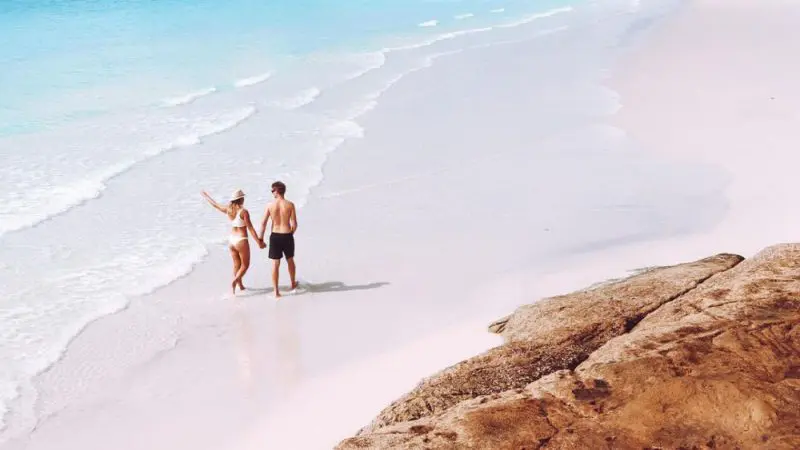 Romantic couple holding hands strolls along a flawless white sand beach beside crystal-clear turquoise water under a sunny blue sky.