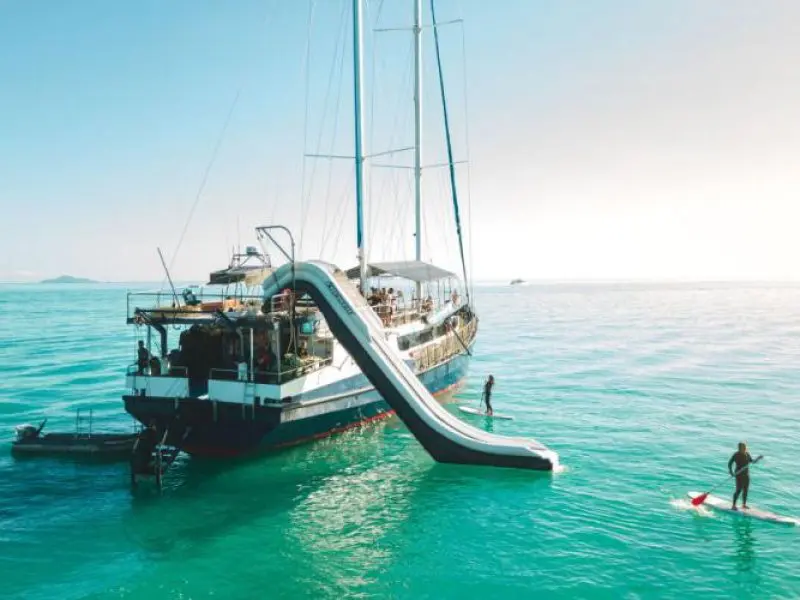 Luxury yacht with slide anchored in crystal-clear blue water; Peterpans Adventure paddleboarder explores under bright sunny skies.