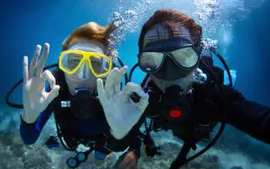 Scuba divers underwater give an OK hand sign in clear blue sea, wearing full wetsuits and masks—perfect diving conditions depicted.