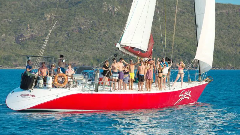 Picture of People wave from a vibrant red sailing boat on clear blue water, with lush green hills rising in the scenic background. This is on a 2 day 1 night social adventure boat called siska.