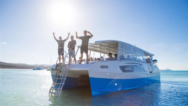 Three smiling travellers wave from the deck of Whitsunday Bullet on a sunny, 1-day Whitsunday Sailing adventure in clear shallow waters.