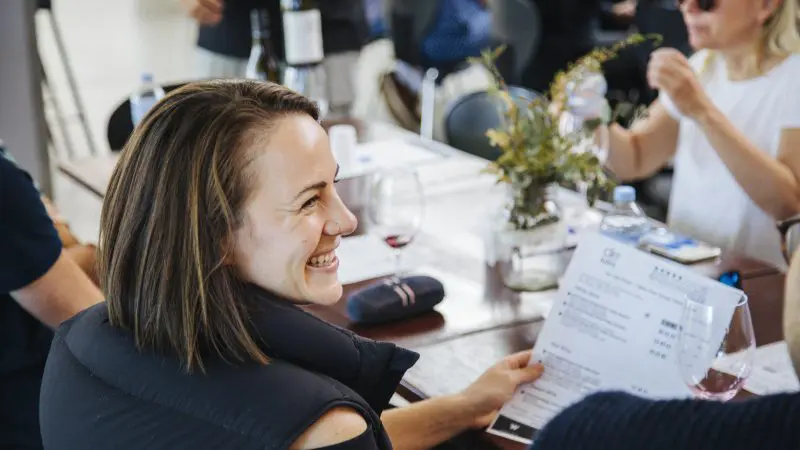 Smiling woman at a sunlit table holding a menu during Colourful Collective’s Hunter Valley wine tour, savouring premium wines.
