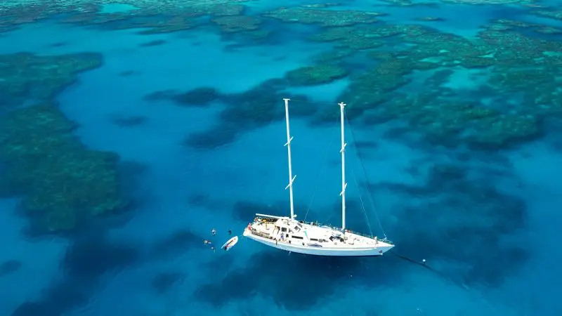 Summer Jo, a white sailboat, anchored in crystal-clear blue waters by vibrant coral reefs during a scenic Whitsunday Sailing tour.