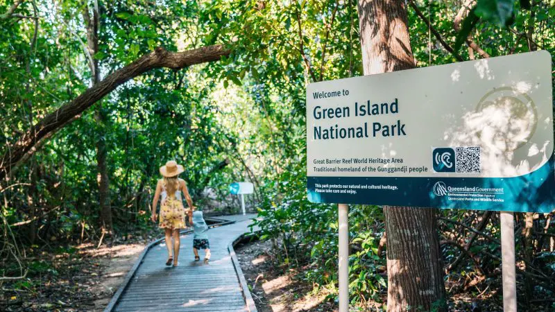 A woman and child stroll along a wooden boardwalk by a Green Island Transfers sign, with lush tropical trees creating a scenic backdrop.