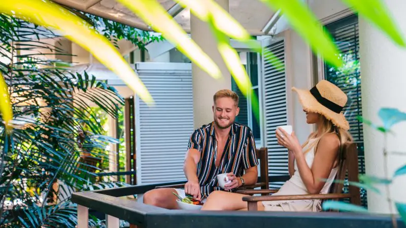 A couple lounges on a lush, leafy patio, sharing memories of snorkelling and glass-bottom boat tours at tropical Green Island.