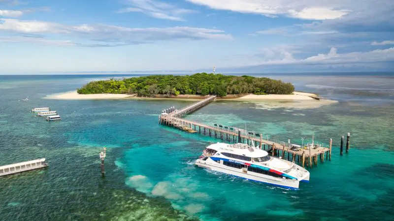 A boat at the Green Island Transfers pier arrives at a tropical island with turquoise waters, coral reefs, and lush green vegetation.