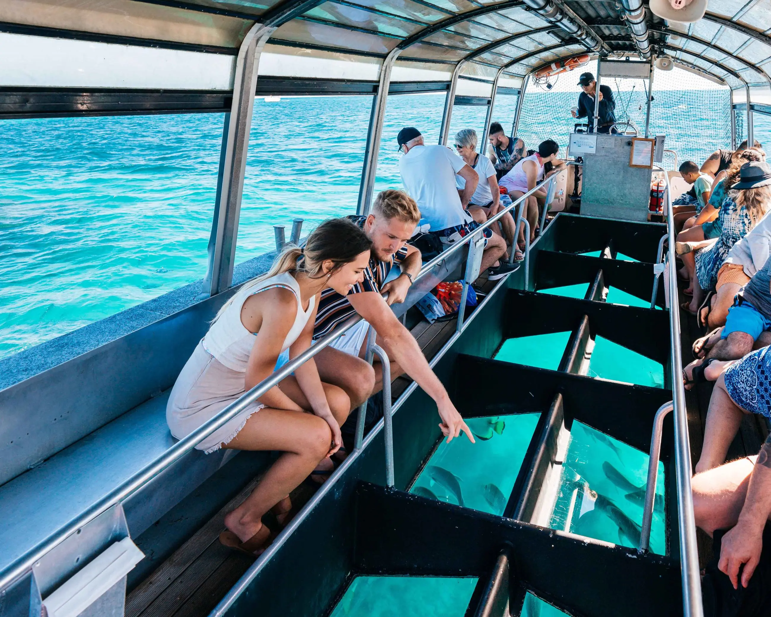 Tourists aboard a glass-bottomed boat observe vibrant tropical fish swimming in crystal-clear blue water beneath the transparent floor.