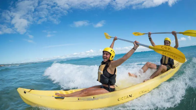 Smiling kayakers in helmets and life jackets paddle on a guided sea tour, raising paddles under a clear blue sky, adventure travel.