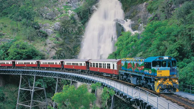 Vibrant Kuranda Scenic Railway train glides over a historic bridge near cascading waterfall and lush, rocky rainforest hills in Australia.
