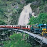 Kuranda Scenic Railway’s colourful Kuranda Deluxe train crosses a bridge near a waterfall and vibrant, lush green rocky hills in Australia.