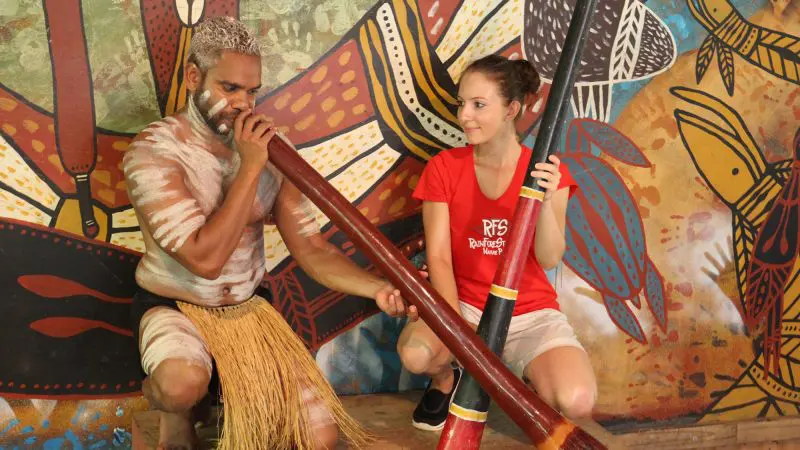 Indigenous Australian man expertly plays didgeridoo as a woman observes during the Kuranda Deluxe & Scenic Railway tour experience.