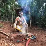 An Indigenous man in vibrant traditional attire sits by a fire, as featured on Express Kuranda Later Departure cultural tour experiences.