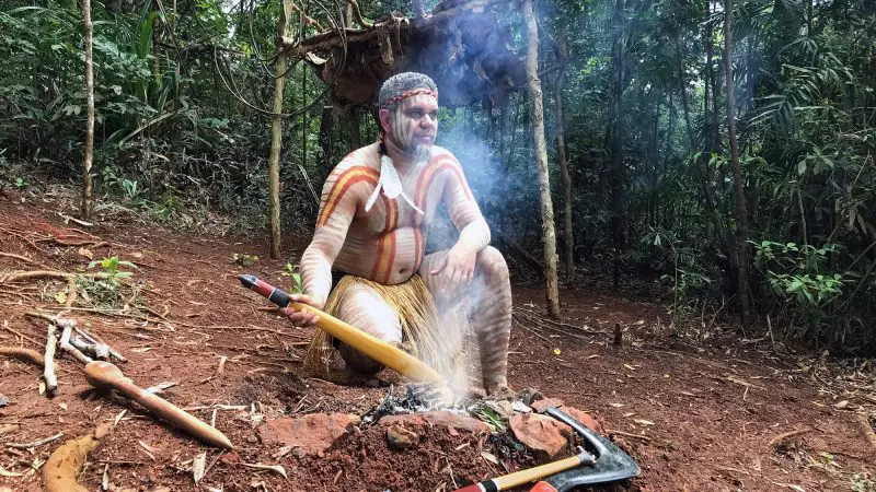 A person in traditional dress sits beside a smoky campfire as the Kuranda Scenic Railway train winds through lush rainforest scenery.