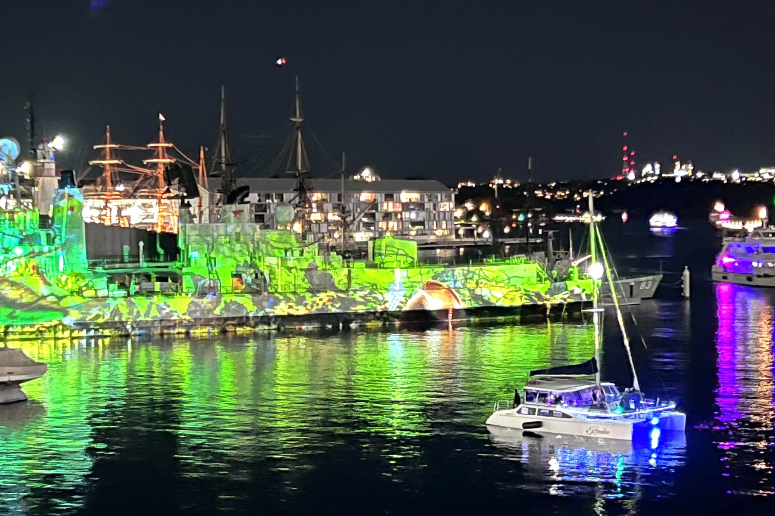 Sydney Harbour illuminated at night with boats and a vibrant Vivid Lights Catamaran Cruise glowing green and purple on the water.