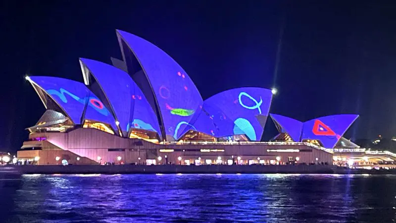 Iconic Sydney Opera House illuminated by vibrant Vivid Sydney lights, glowing and mirrored in the harbour on a BYO catamaran cruise.