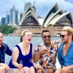Group of friends laughing aboard a catamaran cruise with iconic Sydney Opera House and harbour landmarks in the scenic background.