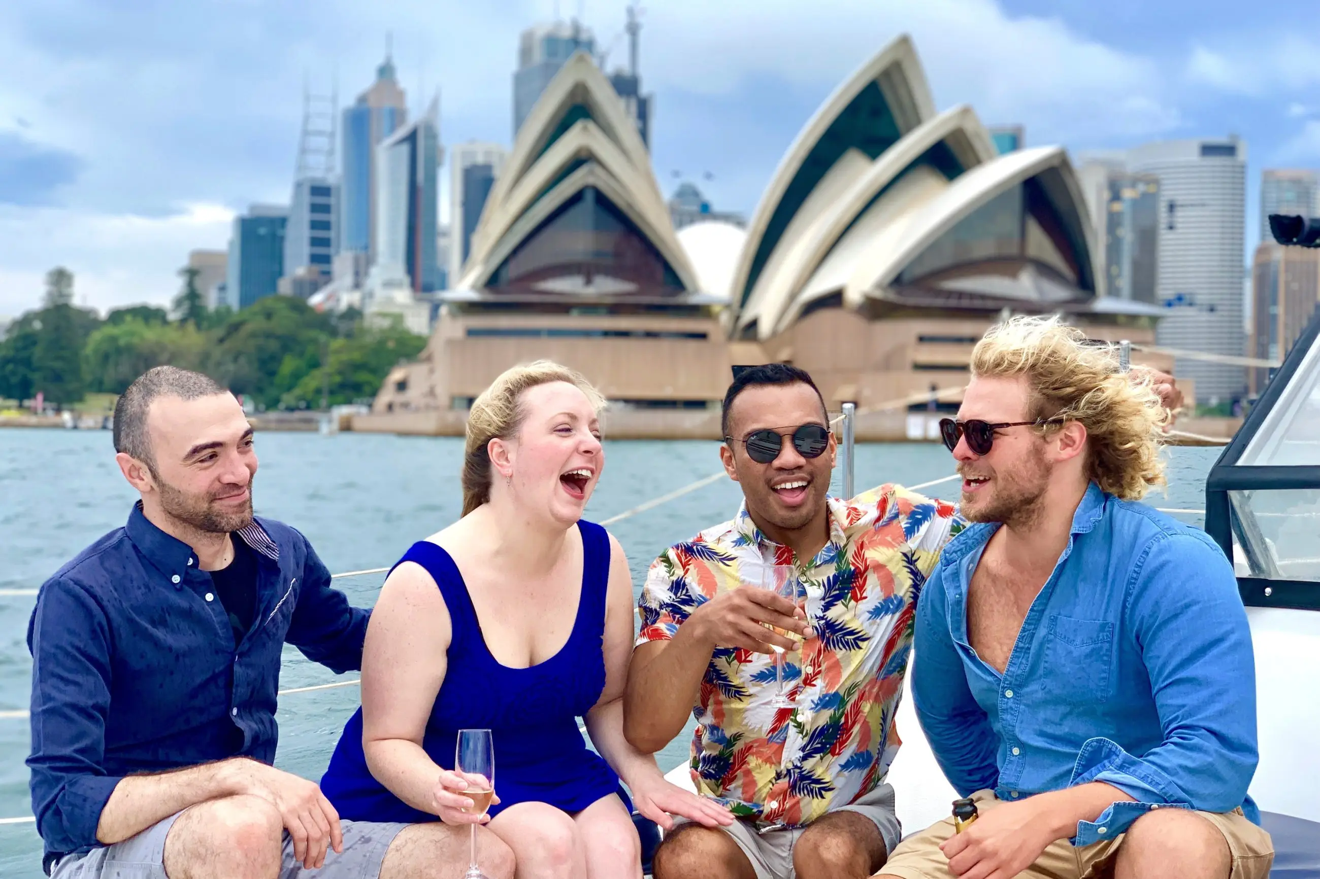 Group of friends laughing aboard a catamaran cruise with iconic Sydney Opera House and harbour landmarks in the scenic background.