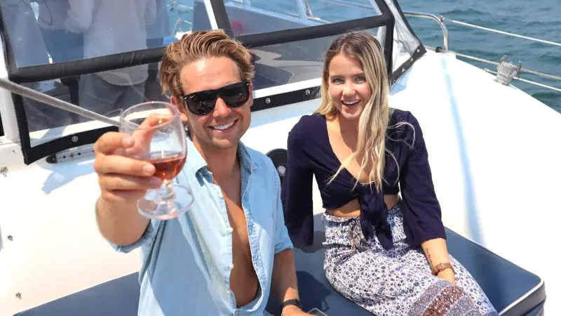 Two happy friends toast with wine on a catamaran, enjoying stunning Sydney Harbour views and sparkling blue water in the background.