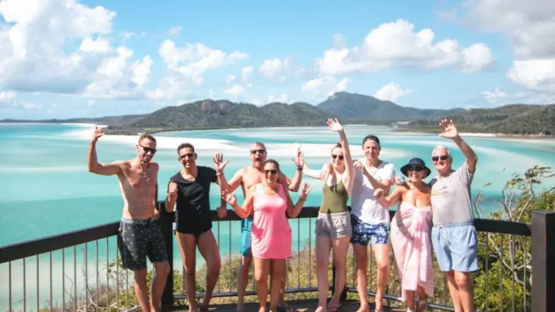 Smiling group in vibrant summer outfits waves from a scenic wooden decking above crystal-clear turquoise Whitsunday waters on holiday.