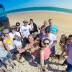 Tour group posing and smiling on Rainbow Beach during K'gari Day Tour with a black SUV, clear blue sky, bright sunny weather.